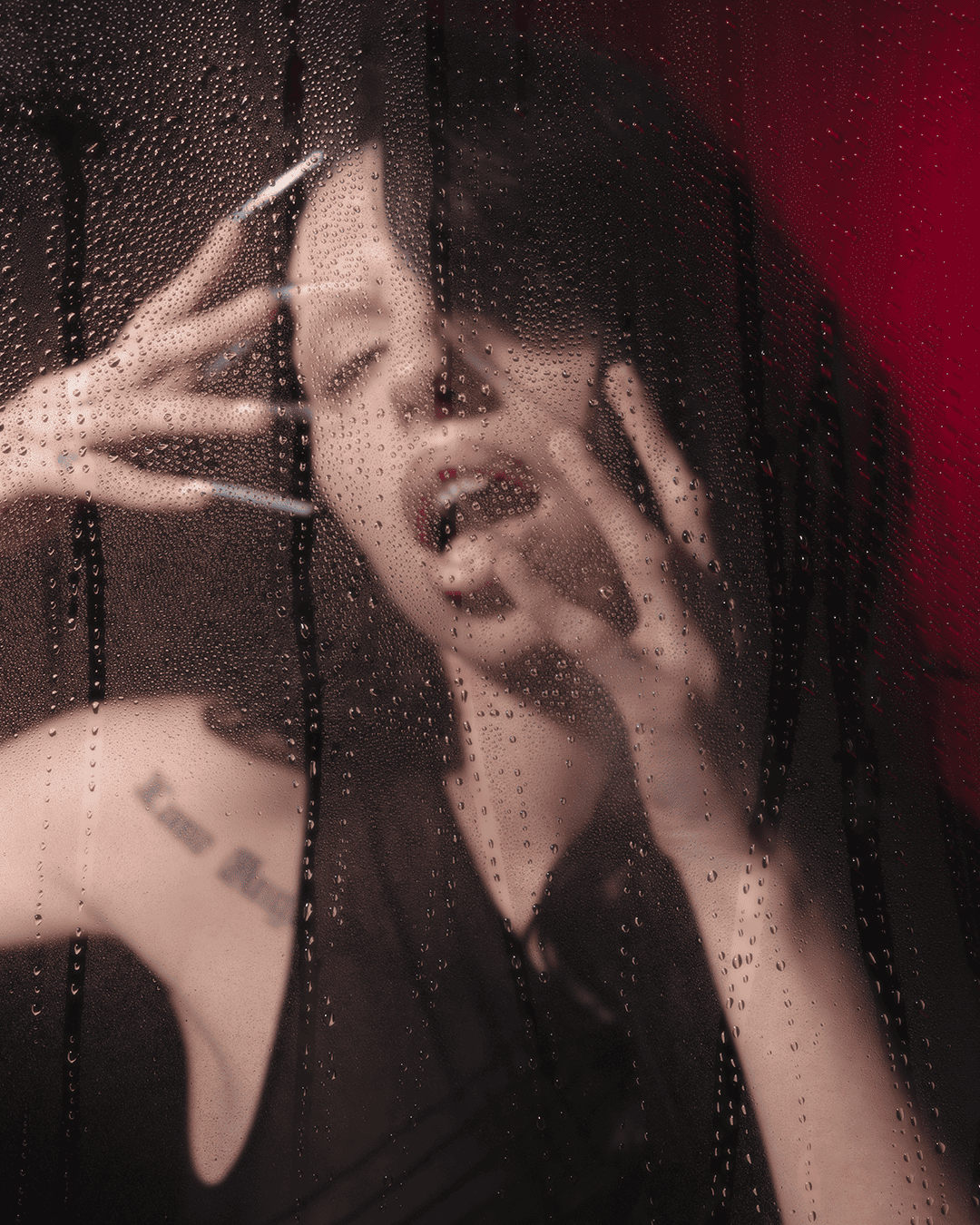 A woman with long nails poses dramatically behind a rain-streaked glass pane, with her mouth slightly open and hands pressed against the glass. The background is dark with hints of red light.