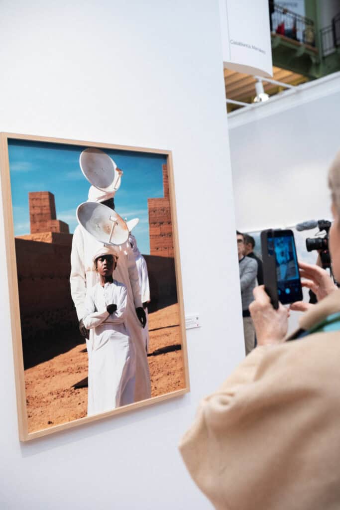 A person photographs a framed artwork by Hiba Baddou in a gallery. The piece shows a person in white before another figure whose head is obscured by two large satellite dishes, set against an outdoor, reddish landscape.