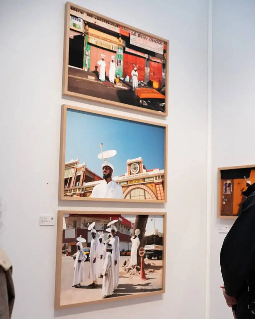 Three framed photographs of people dressed in white are displayed on a white gallery wall; two people partially visible view the artwork. The top and bottom photos feature groups, the middle has a single person.
