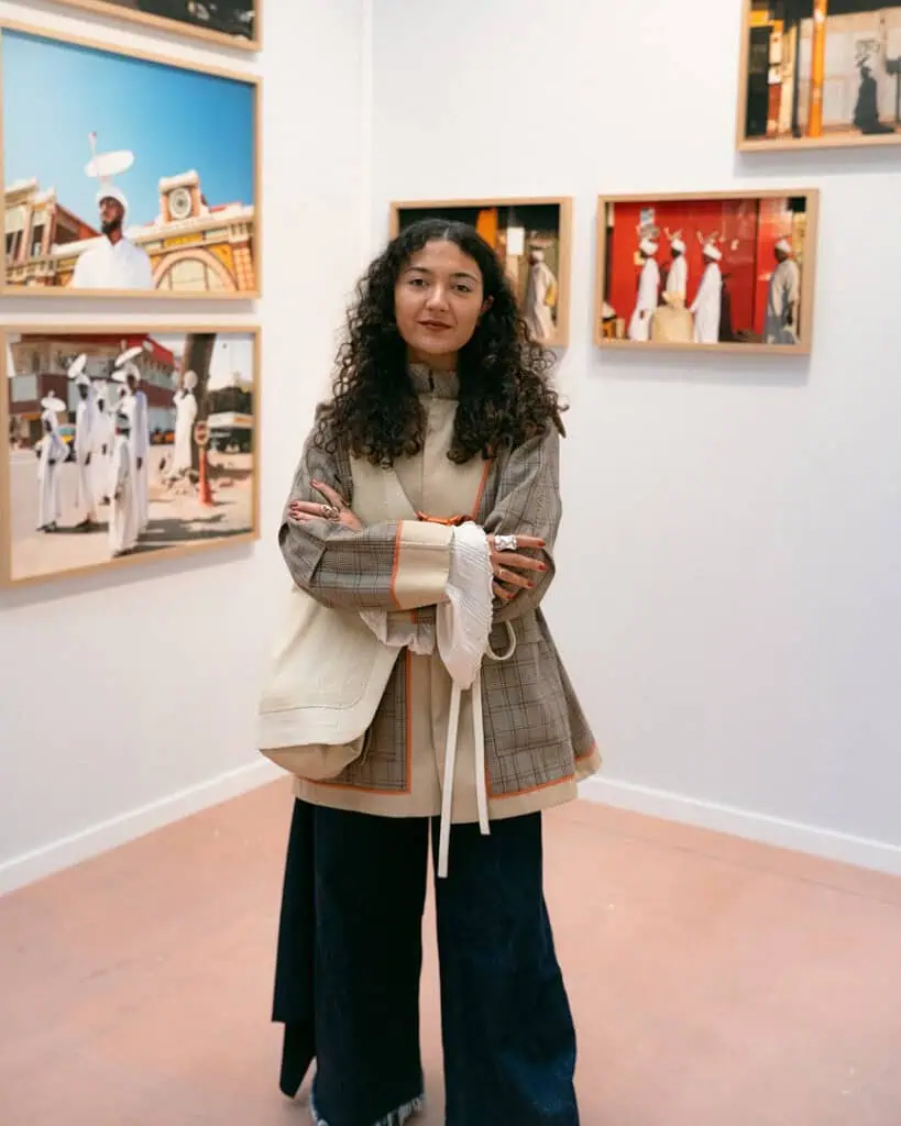 A woman with long curly hair stands confidently with arms crossed in an art gallery, wearing layered, stylish clothing. Framed photographs of people dressed in traditional attire are displayed on the walls behind her.