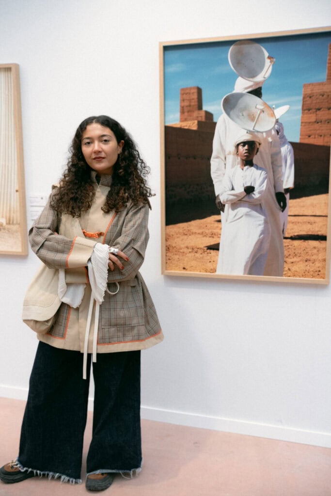 A woman with curly hair stands indoors next to a framed photograph by Hiba Baddou, featuring people in white clothing and large headpieces against a desert-like backdrop. She wears a layered, patterned outfit and dark wide-leg jeans.
