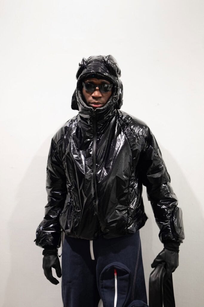 A person wearing a shiny black hooded jacket, black gloves, and dark sunglasses stands against a plain white background. Their outfit has a futuristic or protective look.