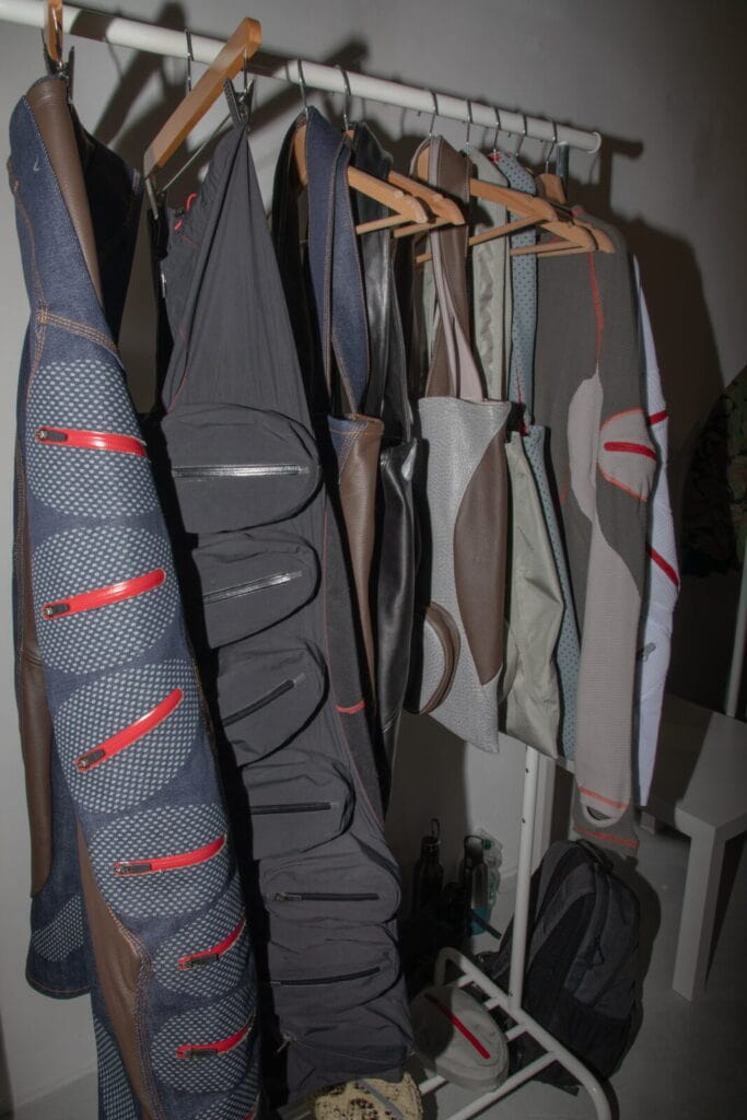 A clothing rack displays several uniquely designed jackets and garments with zippers and geometric patterns in shades of gray, blue, brown, and red. A backpack and other items sit on the floor nearby.