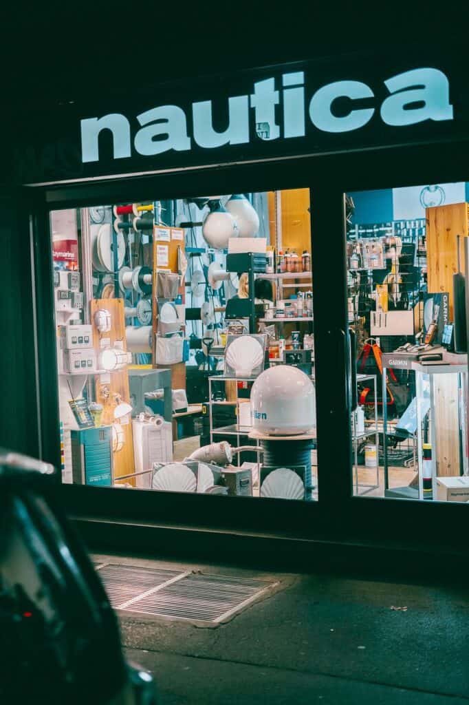 A store window at night displays various nautical equipment and supplies under a bright nautica sign. The interior is filled with lights, gadgets, and marine-related items.