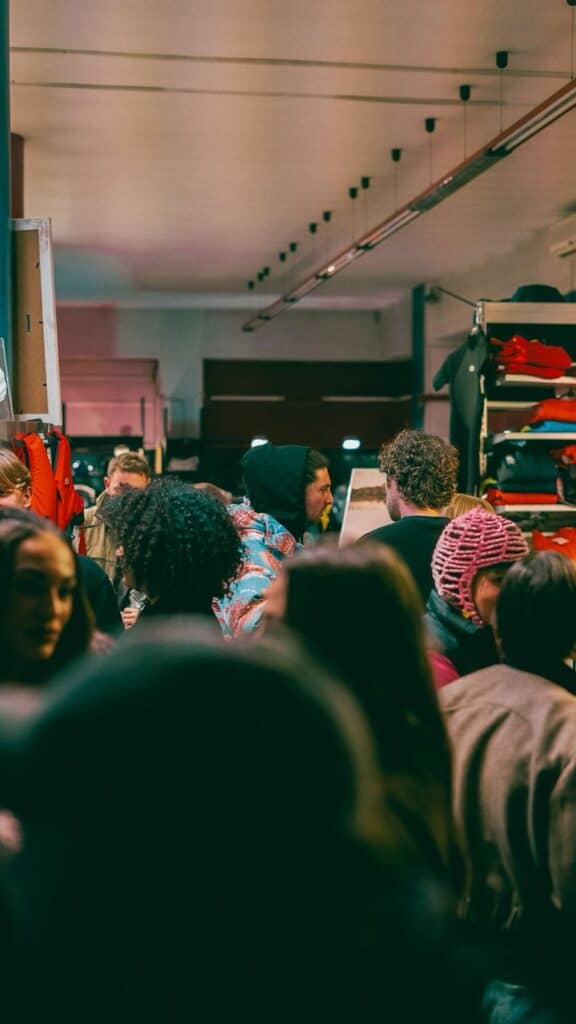 A crowded indoor space with people talking and mingling. Some wear winter clothing like beanies and coats. The background shows shelves with red and orange clothes on display. The lighting is warm and ambient.