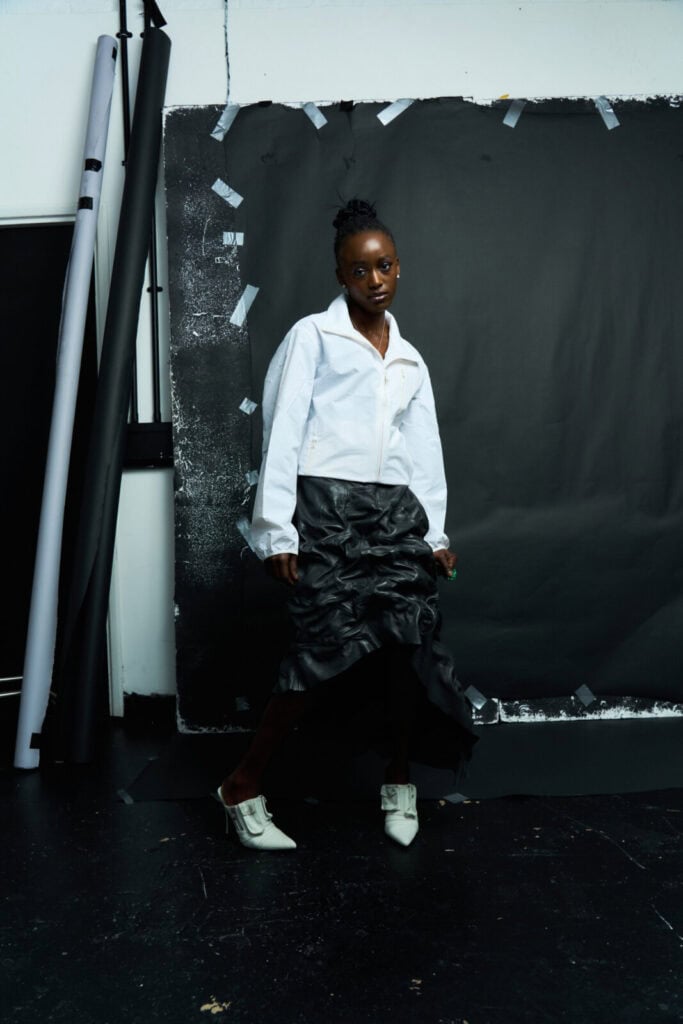 A person poses indoors against a black backdrop, wearing a white collared shirt, a black ruffled skirt, and white pointed shoes. The setting appears artistic, with rolls of paper and tape visible around the backdrop.