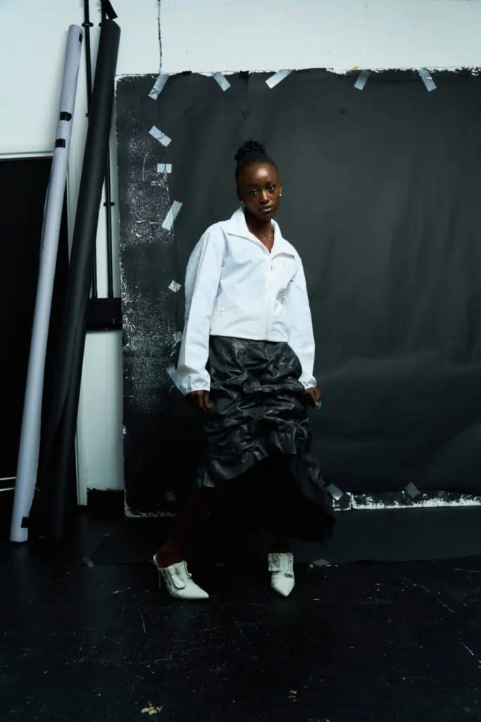 A person poses indoors against a black backdrop, wearing a white collared shirt, a black ruffled skirt, and white pointed shoes. The setting appears artistic, with rolls of paper and tape visible around the backdrop.