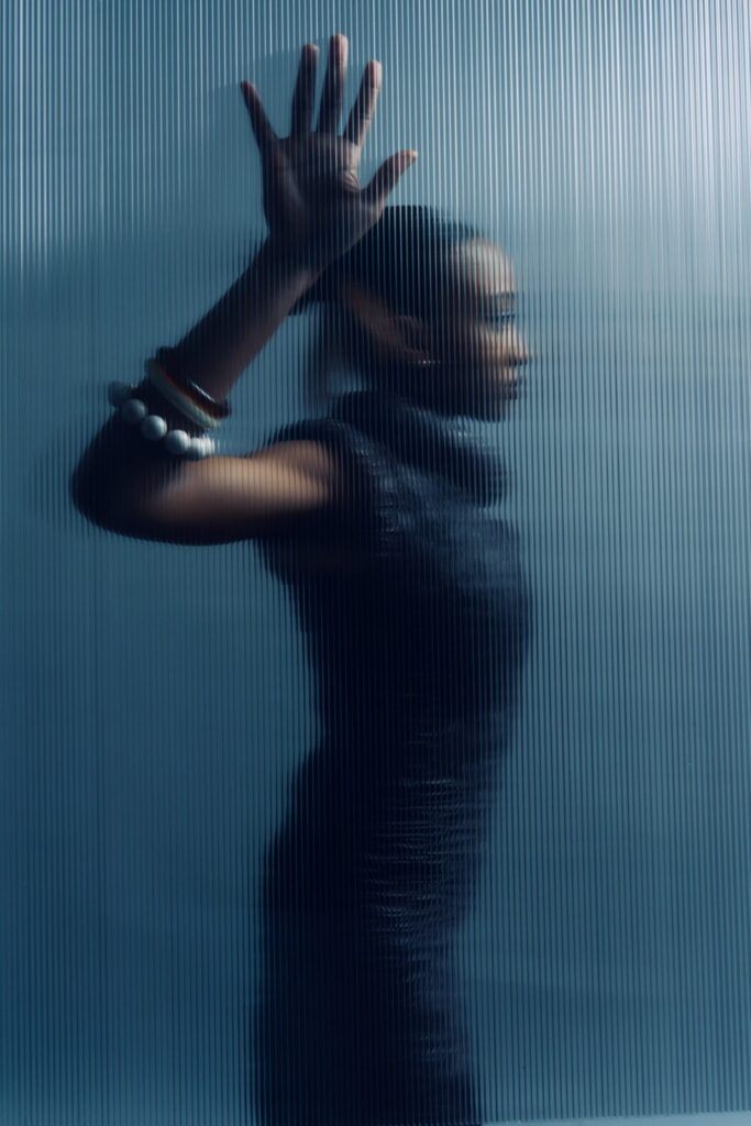 A woman stands behind a frosted, ribbed glass panel with her hand pressed against it. Her profile and features are blurred, and she wears bracelets and a dark sleeveless dress. The background and lighting are cool-toned.