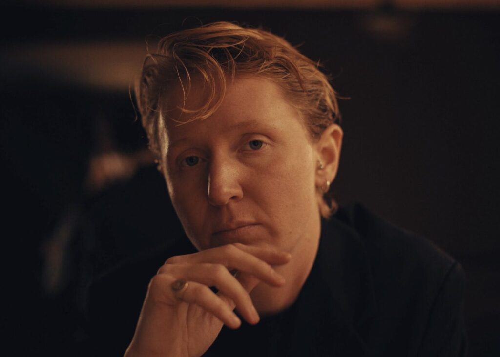 A person with short, wavy reddish-blonde hair and light skin looks thoughtfully at the camera, resting their hand near their chin in a dimly lit setting.