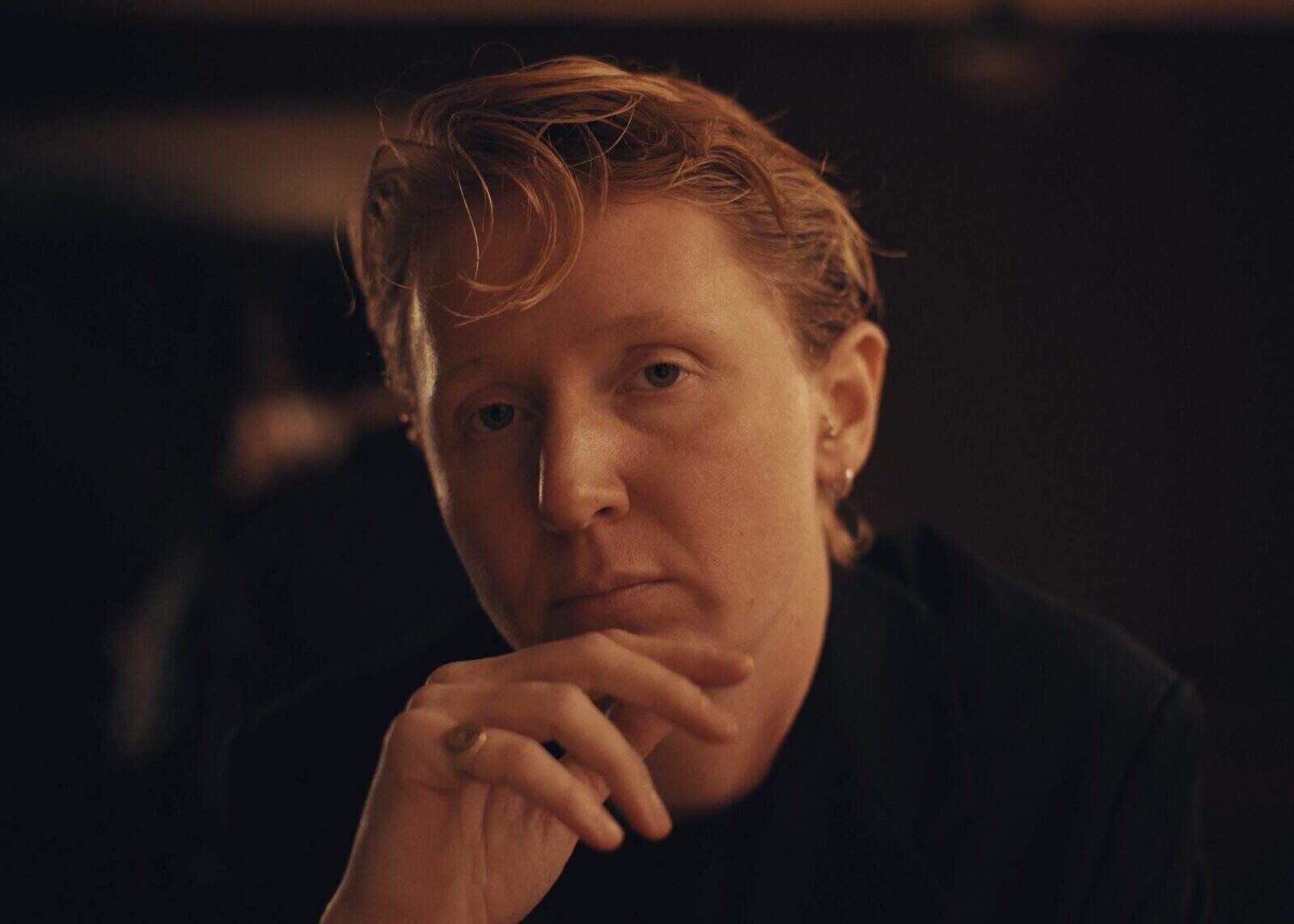 A person with short, wavy reddish-blonde hair and light skin looks thoughtfully at the camera, resting their hand near their chin in a dimly lit setting.