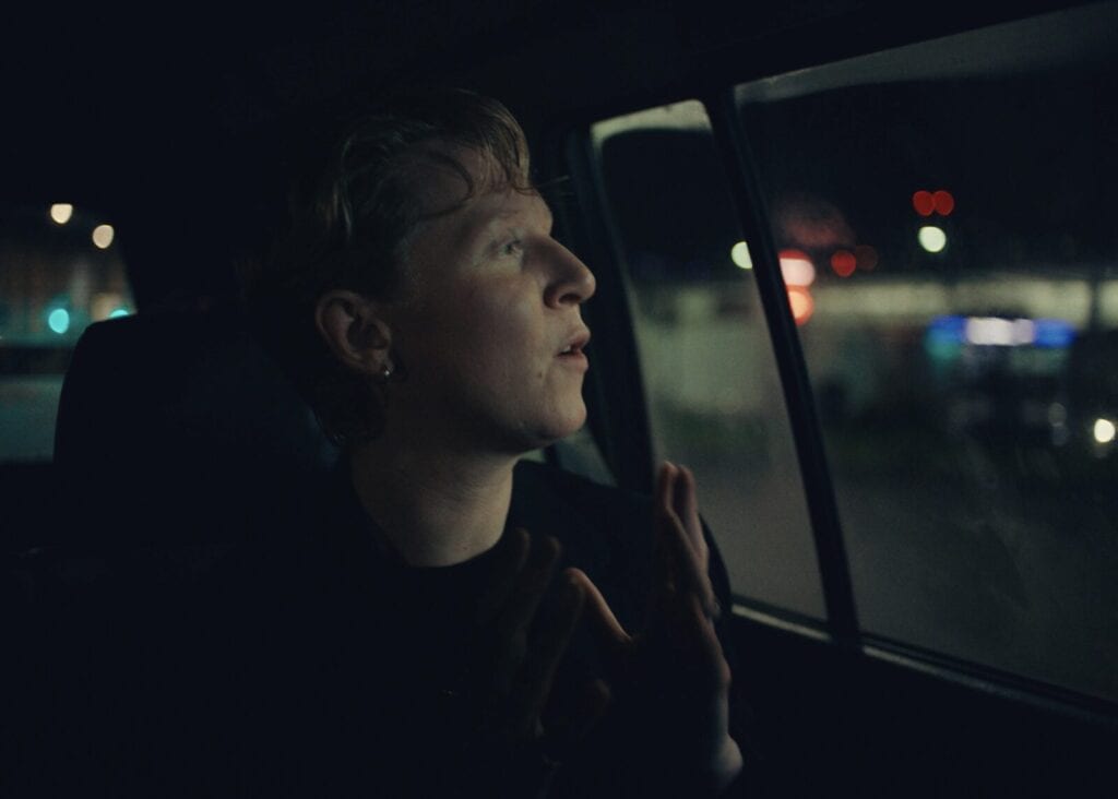A person with short hair sits in a dimly lit car at night, looking out the window with hands raised near their face. City lights and blurred night scenery are visible outside.