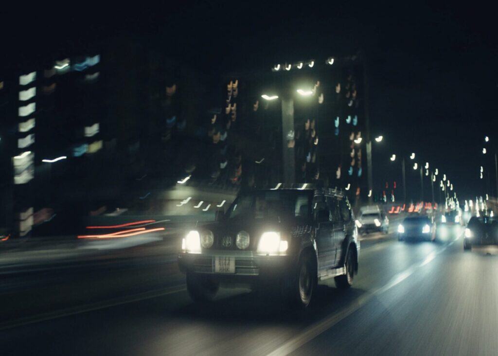 A blurred night-time scene of cars driving quickly down a city street, with headlights shining and city lights reflecting off buildings in the background.