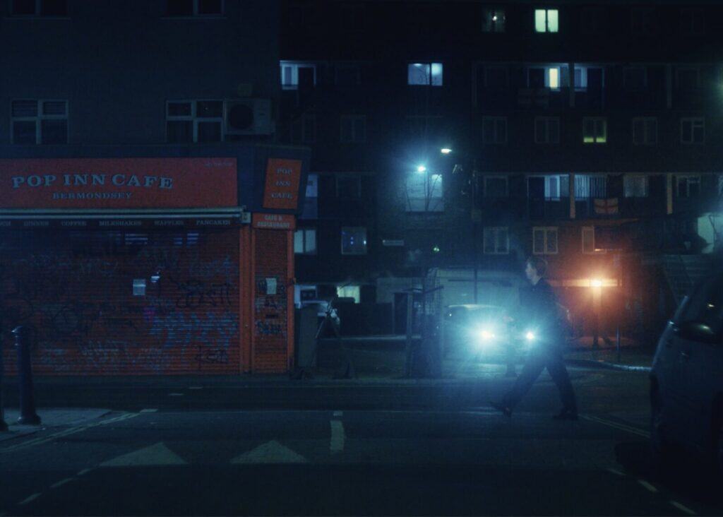 A person crosses a dimly lit street at night in front of the closed, red-brick Pop Inn Cafe, with glowing windows and headlights from a car in the background.
