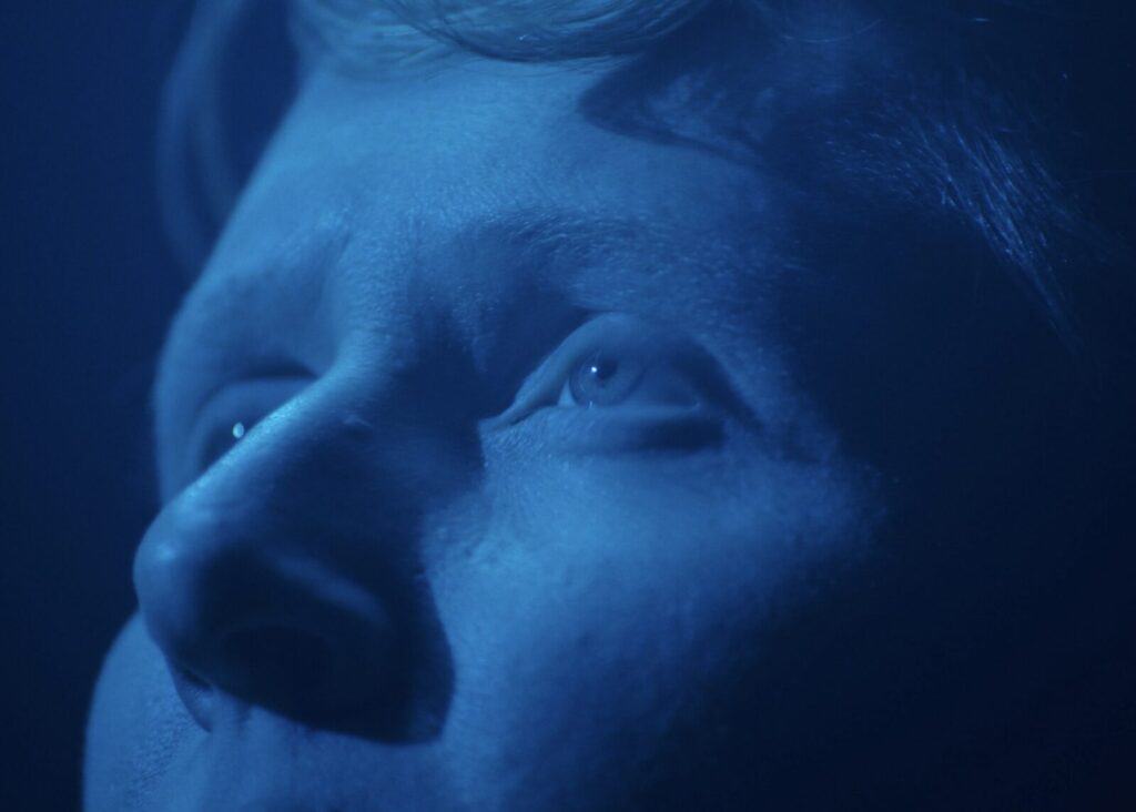 A close-up of a persons face lit with blue light, focusing on their eyes looking upward with a thoughtful expression.