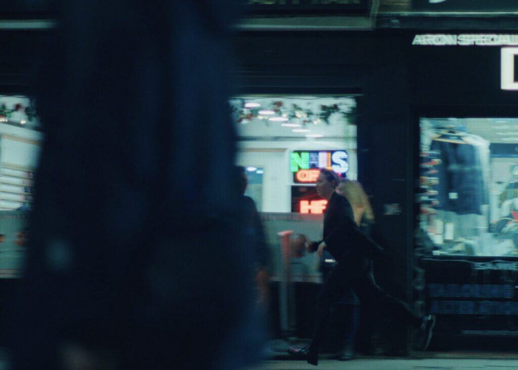 A person in a suit is running on a city street at night, passing by a nail salon with neon signs. The image is slightly blurred, suggesting motion, and another figure is partially visible in the foreground.