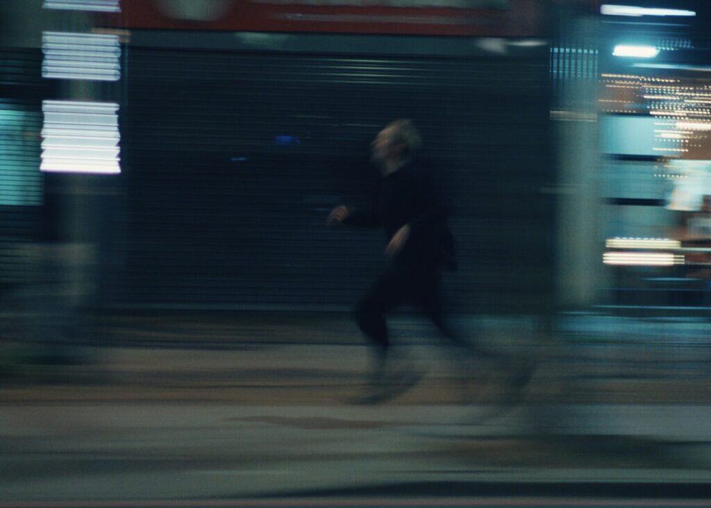 A blurred image of a person running quickly on a city street at night, with bright lights and storefronts in the background. The motion creates a sense of urgency and speed.