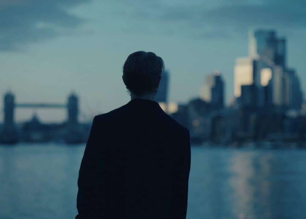 A person with short hair, dressed in dark clothing, stands by a body of water looking toward a city skyline at dusk. The scene includes blurred skyscrapers and a faint outline of a bridge in the background.