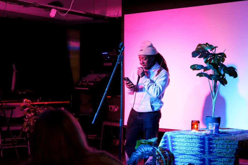 A person in a white hoodie reads from their phone while speaking into a microphone on stage, lit by pink and blue lights. A Valentine-themed plant and a lit candle sit on a decorated table nearby.
