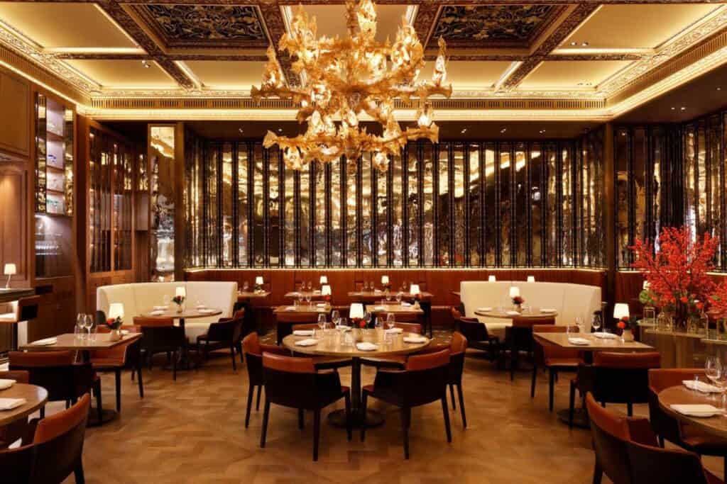 Elegant restaurant interior with round tables, white booths, brown chairs, a grand golden chandelier, warm lighting, wooden floors, and decorative gold accents. Red flowers and table settings add a refined touch, advice about where to eat during London Fashion week.