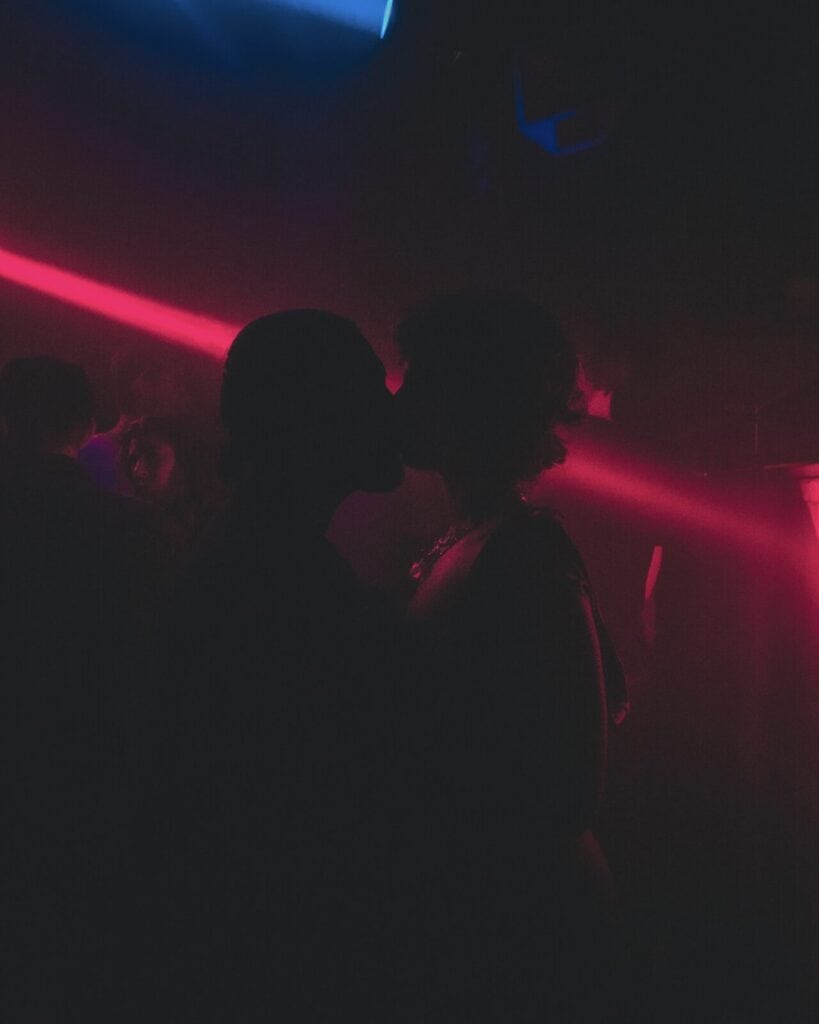 Two people are silhouetted as they kiss in a dimly lit room with red and blue neon lights and smoky atmosphere, surrounded by other faintly visible figures in the background.