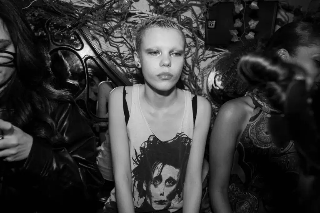 A person with short hair and bleached eyebrows sits between two others in a crowded room, wearing a tank top with a graphic of Edward Scissorhands. The black-and-white photo captures the buzz, perfect for those seeking Where To Eat During London Fashion Week.