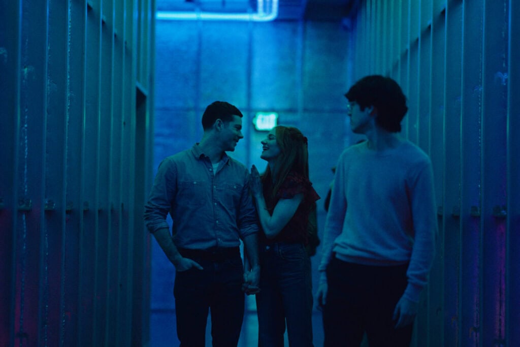 Three people walk through a dimly lit hallway with blue lighting. A couple, holding hands and smiling at each other, walk together while a third person walks slightly ahead, looking back at them.