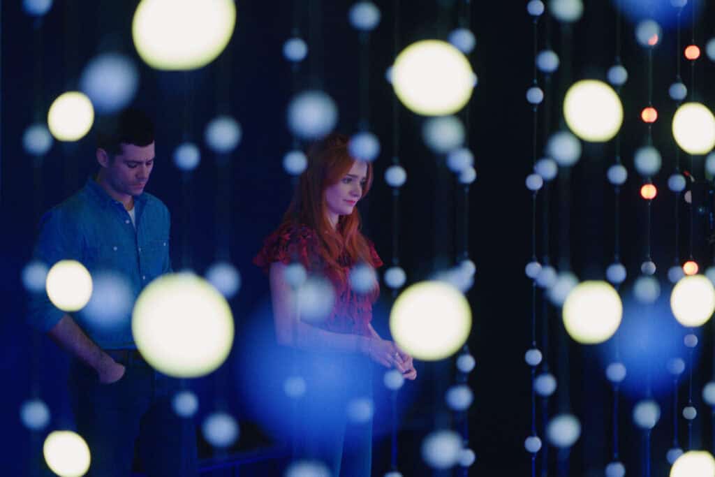 A man and a woman stand in a dimly lit room, partially obscured by hanging strings of glowing orbs. The background is dark, highlighting the illuminated orbs and creating a dreamy, contemplative atmosphere.