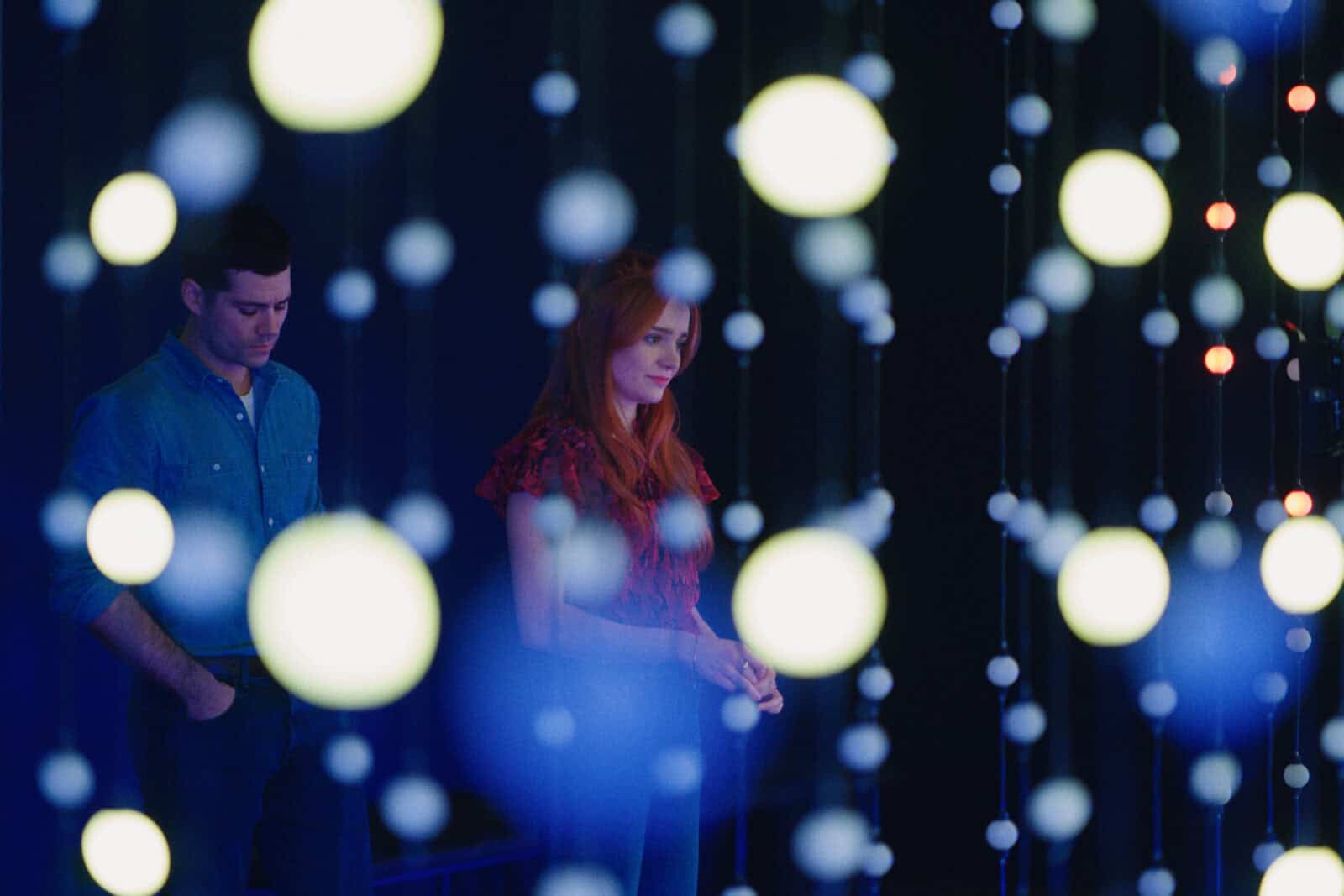 A man and a woman stand in a dimly lit room, partially obscured by hanging strings of glowing orbs. The background is dark, highlighting the illuminated orbs and creating a dreamy, contemplative atmosphere.