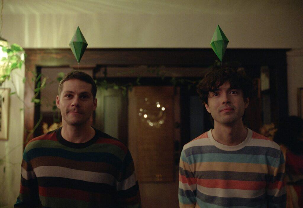 Two men stand side by side indoors, each wearing a headband with a green plumbob above their heads, referencing The Sims video game. They both wear striped sweaters and look forward with neutral expressions.