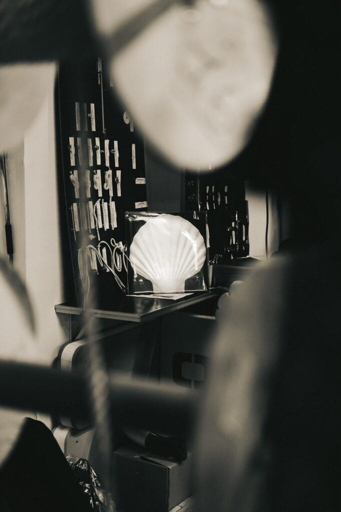 A black and white photo showing a room with a glowing shell-shaped lamp on a desk, partially obscured by blurred objects in the foreground. Various items and tools hang on the wall behind the desk.