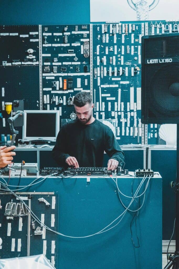 A man stands behind a DJ mixer, adjusting controls. He is surrounded by wires and electronic equipment, with a wall covered in tools and devices in the background. A large speaker is visible on the right.