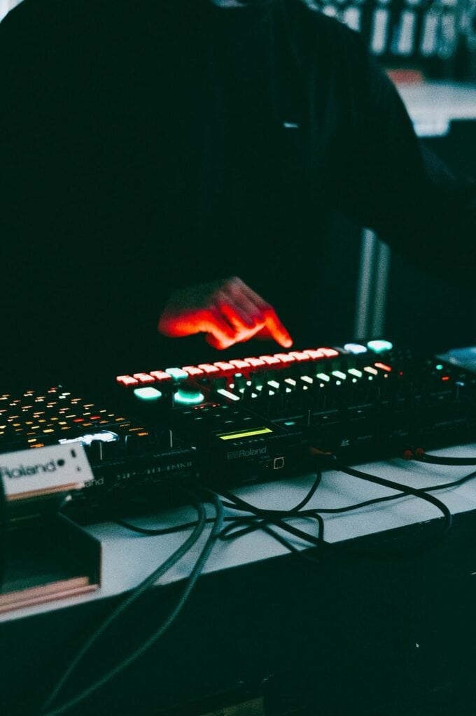 A person in dark clothing presses buttons on an illuminated DJ mixing console, surrounded by cables and electronic equipment in a dimly lit setting.