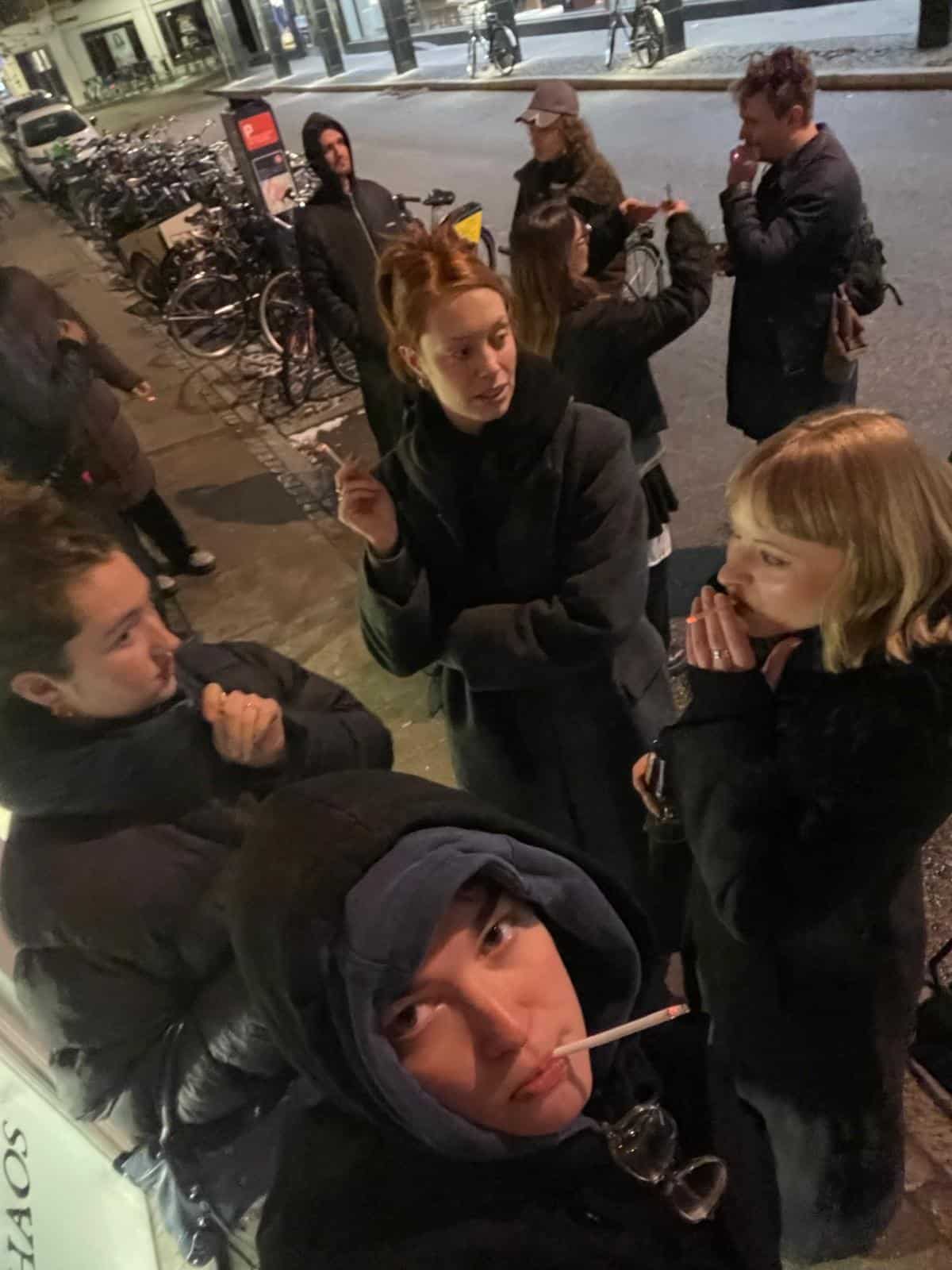 A group of young people stand outside at night in heavy coats. Some are smoking and talking, while bicycles are parked along the sidewalk behind them. The scene appears casual and social.