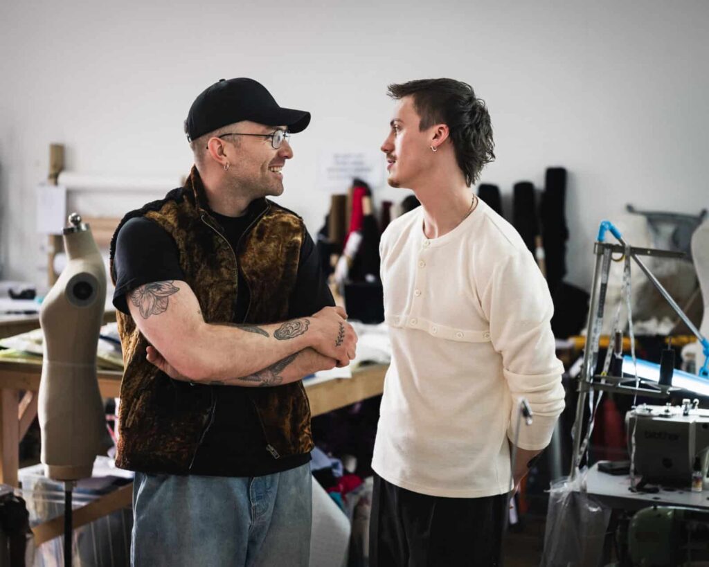 Agro studio founders stand and talk in a workshop or studio. One wears a black cap, glasses, and a brown vest with visible tattoos on his arms; the other wears a white shirt. Sewing equipment and mannequins are in the background.