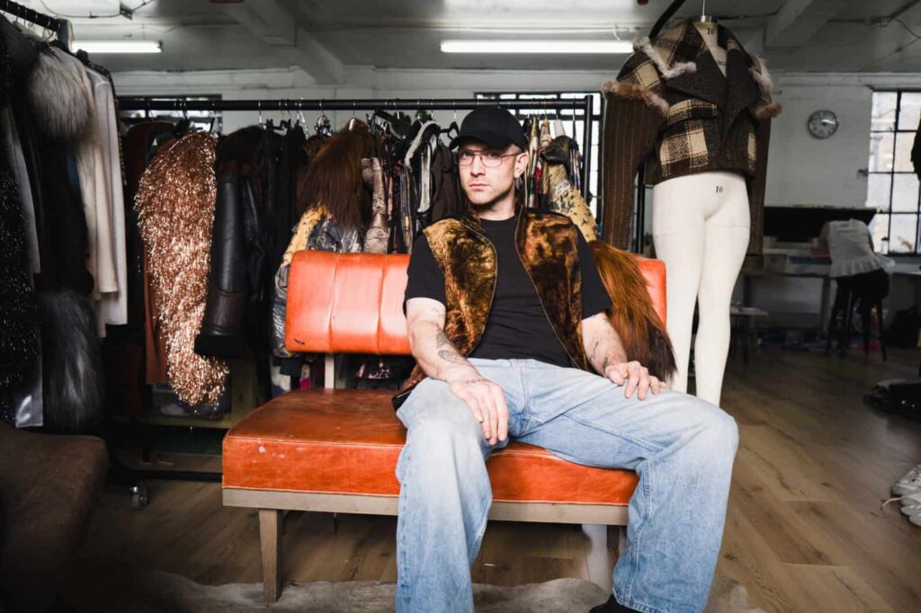 Agro studio founder a fur vest, black cap, and glasses sits on an orange sofa in a studio with clothing racks and a dress form behind him. The space has wooden floors and large windows.