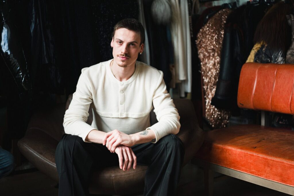 The founder with short dark hair and a mustache sits on a brown chair, wearing a white buttoned sweater and black pants. Clothes on hangers and an orange bench are in the background.