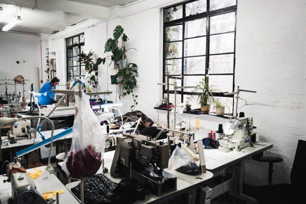 Bright sewing studio with several sewing machines on tables, potted plants on windowsills, and two people working in the background near large windows letting in natural light.