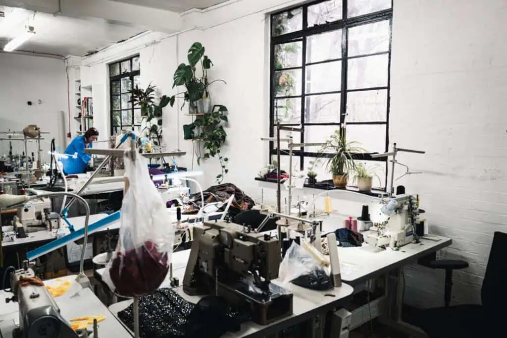 Bright sewing studio with several sewing machines on tables, potted plants on windowsills, and two people working in the background near large windows letting in natural light.