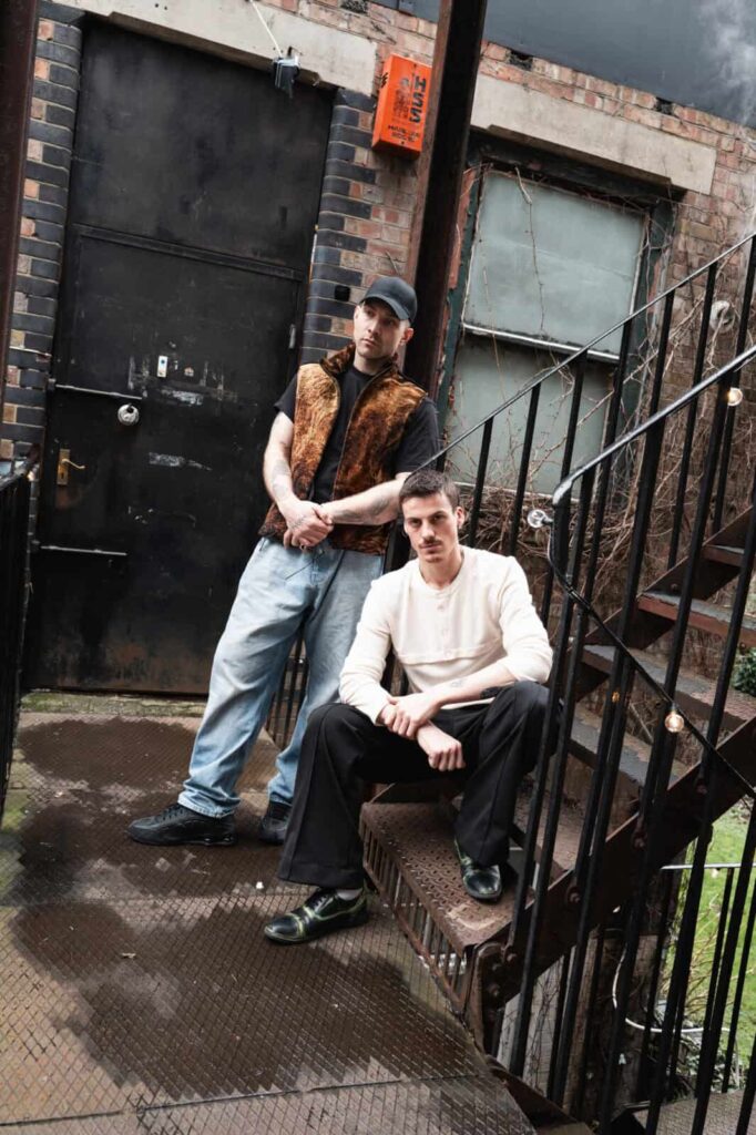 Two men pose on an outdoor metal staircase by a brick building. One stands with arms crossed, wearing a cap, vest, and jeans. The other sits, wearing a white sweater and black pants. The background is gritty and urban.