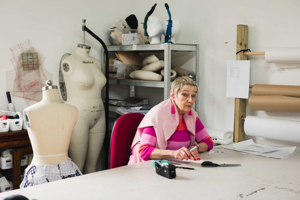 A woman wearing a pink striped sweater sits at a workspace with sewing tools and fabric. Behind her are mannequins, shelves with rolls of material from Agro studio, and various sewing supplies. She looks thoughtfully towards the camera.