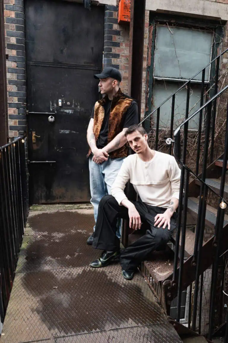 Two men pose on a metal stairway and landing outside a brick building. One stands by a black door, wearing a brown vest and cap, while the other sits on the steps in a white shirt and black pants.