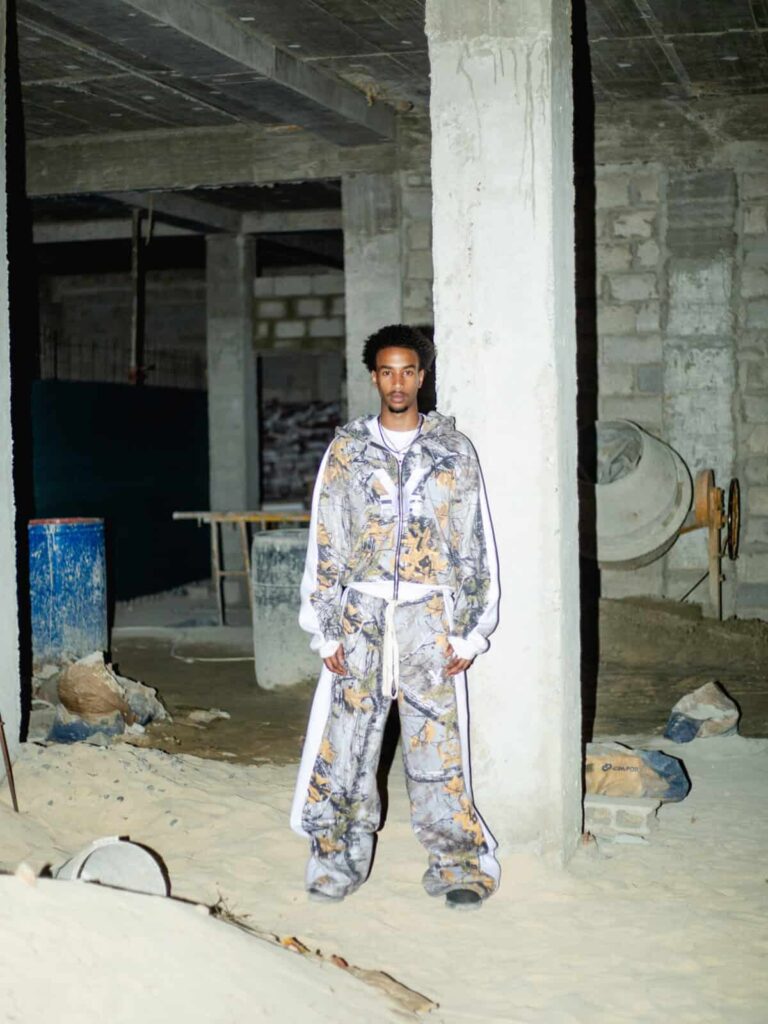 A man wearing a matching All-T¥lk patterned hoodie and baggy pants stands in a dimly lit, unfinished concrete building with sand and construction materials scattered around.