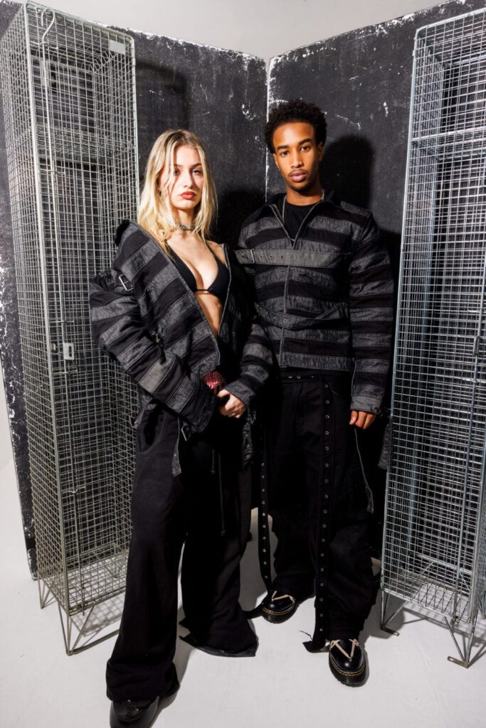 Two models stand side by side in front of wire lockers, wearing All-T¥lk coordinated black outfits with wide horizontal stripes and details. The woman’s jacket is open, revealing a black bra, and both display confident expressions.