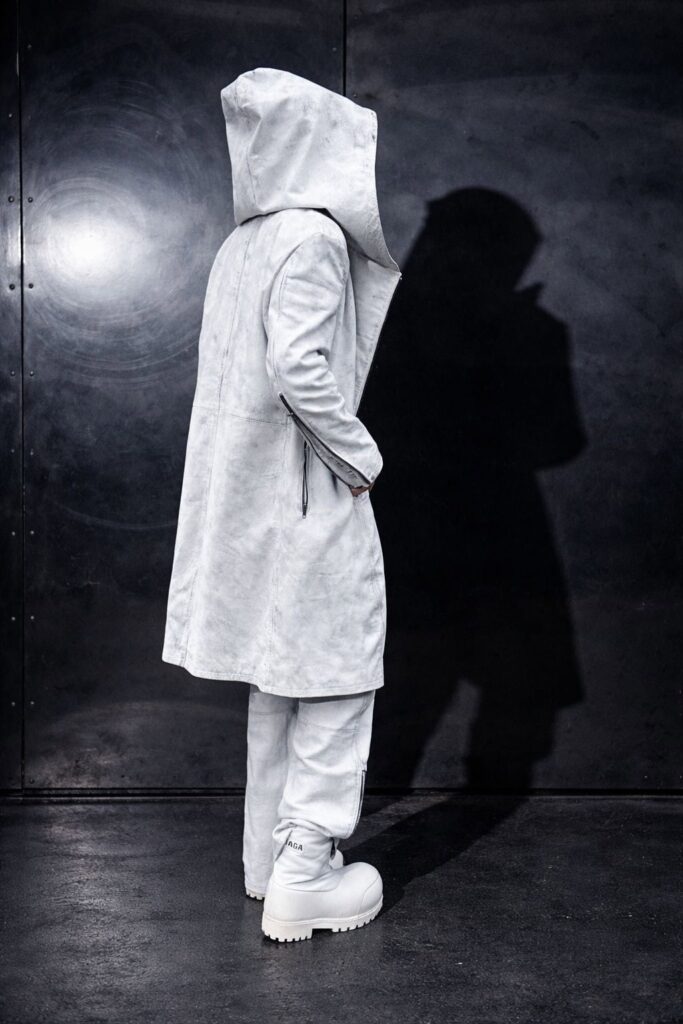 A person in a light gray All-T¥lk hooded coat, matching pants, and chunky white boots stands against a dark wall, facing away with their head turned slightly to the side, casting a shadow on the wall.