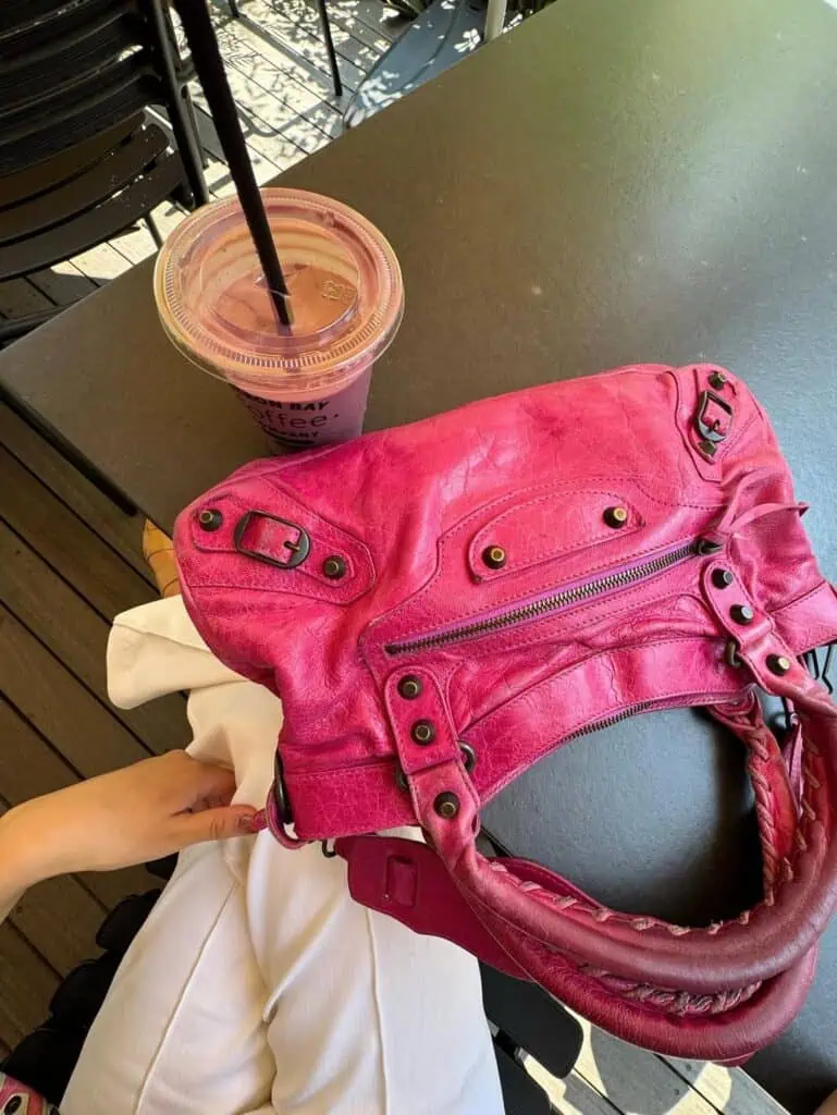 A bright pink HIVE handbag rests on a dark table next to a purple smoothie in a clear cup with a black straw. A person wearing white pants sits nearby, with their hand holding the bag.