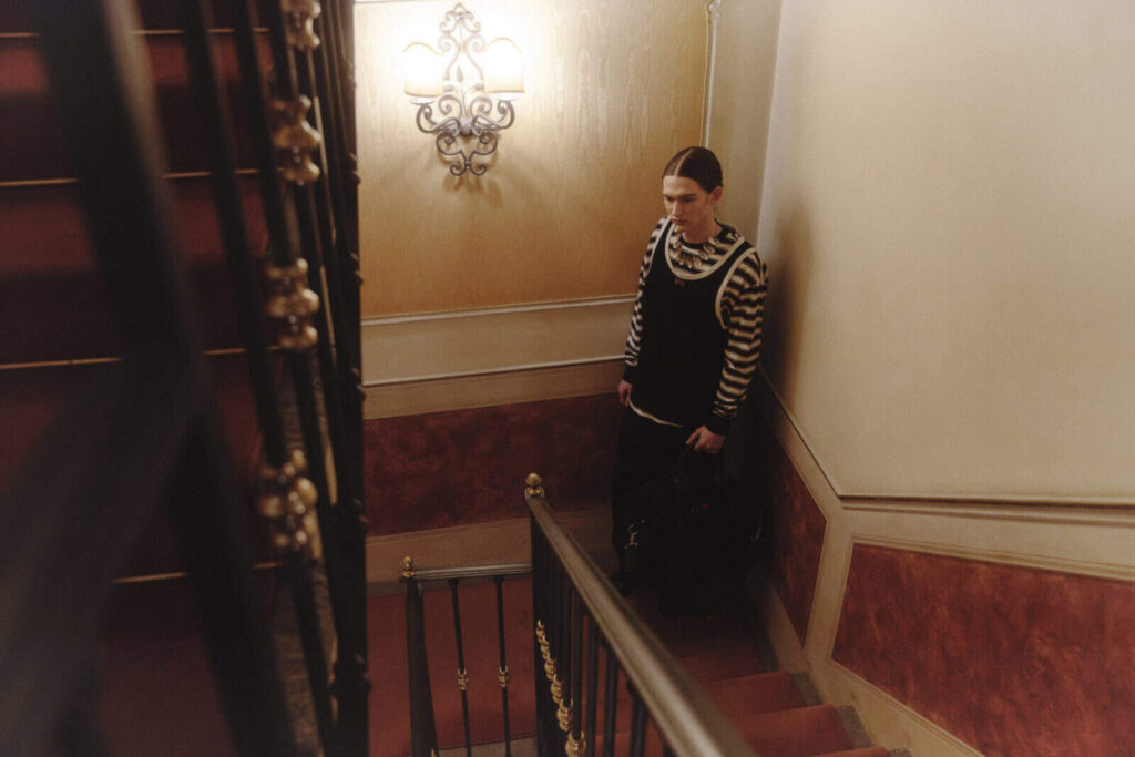A person wearing a black dress over a black-and-white striped shirt stands on a red-carpeted staircase next to an ornate wall lamp in a warmly lit, vintage-style interior.