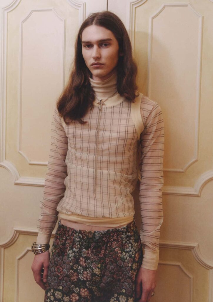 A person with long brown hair stands in front of a cream-colored paneled wall, wearing a sheer, plaid long-sleeve top over a turtleneck and floral-patterned trousers. They have bracelets on one wrist and a neutral expression posing for Before We Could Speak editorial.