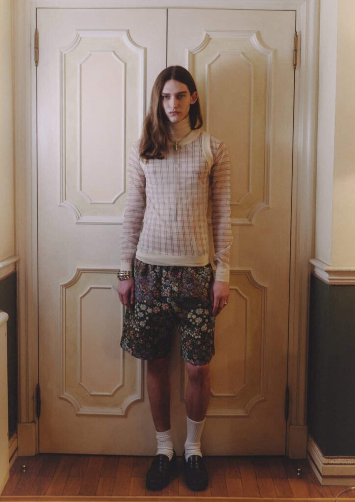 A person with long hair stands indoors against double doors, wearing a sheer, long-sleeved plaid top over a white turtleneck, floral shorts, white socks, black shoes, and several bracelets, looking directly at the camera posing for Before We Could Speak editorial.