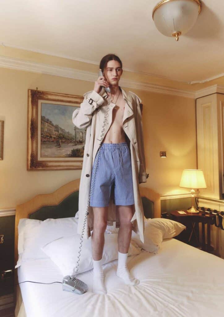 A young person stands on a hotel bed wearing a trench coat, blue striped boxer shorts, white socks, and holding a corded phone to their ear. The room features warm lighting, classic decor, and a framed painting on the wall for Before We Could Speak editorial.