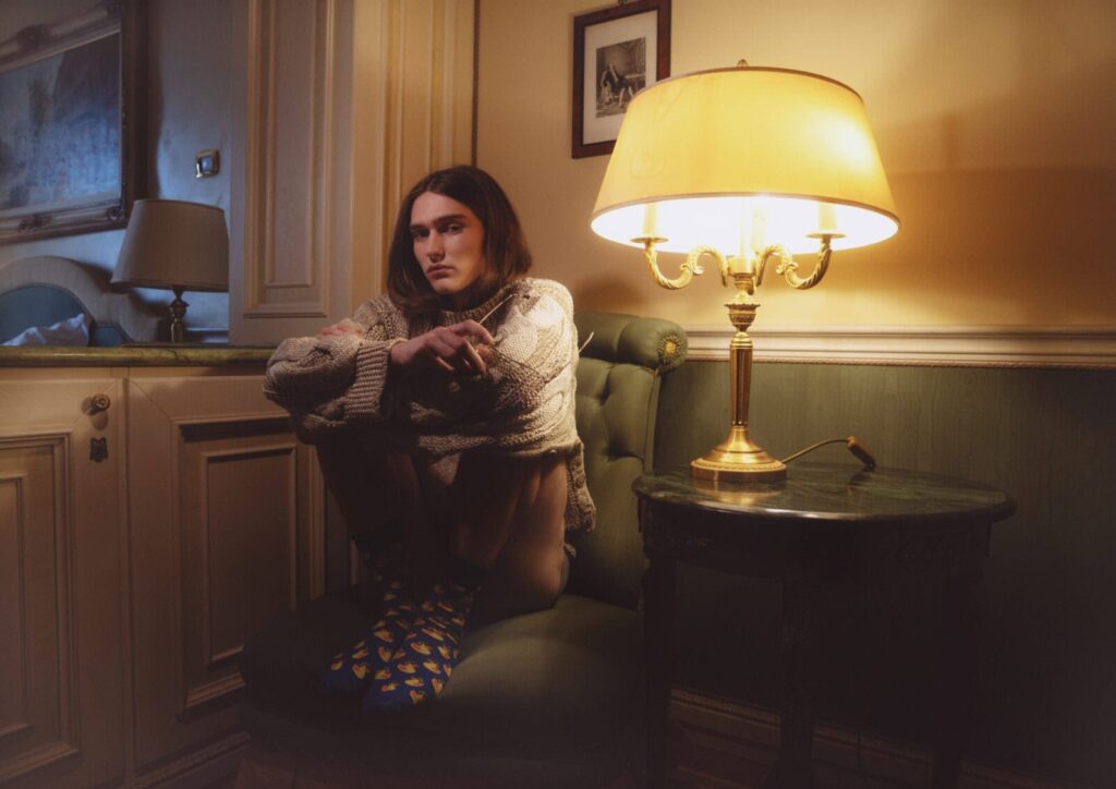 A person with long hair sits barefoot on a green armchair, hugging their knees, wearing a thick sweater and patterned socks. A lit table lamp and framed pictures create a cozy, warm atmosphere in the room posing for Before We Could Speak editorial.